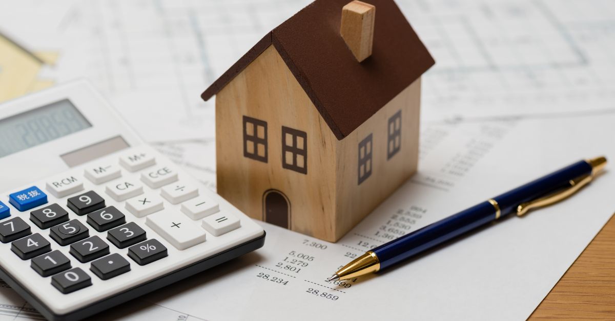 A wooden toy house with a calculator, paper and a pen around it which were used to compute the cost to build a house in Austin.